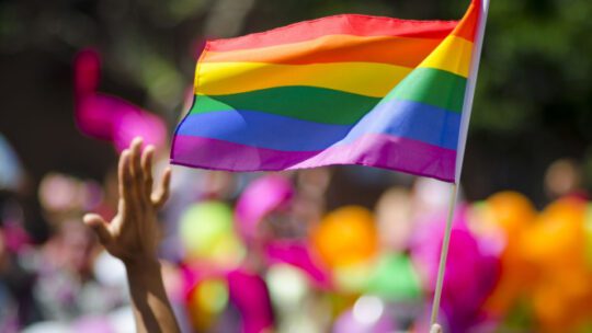 A supporting hand waves in front of a rainbow flag flying on the sidelines of a summer gay pride parade