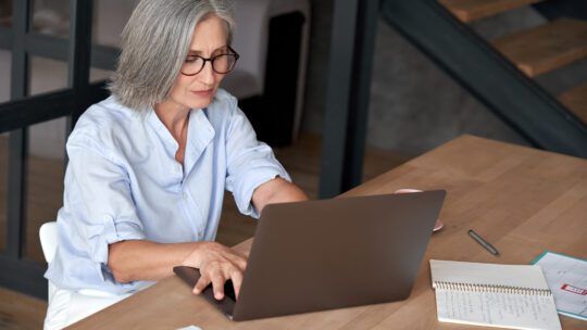 Older mature middle aged business woman employee using laptop typing computer sitting at workplace desk. Senior old lady 60s grey-haired businesswoman entrepreneur or executive working on pc in office