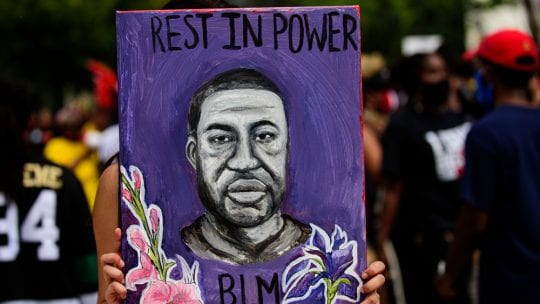 Protestor holds purple poster featuring George Floyd portrait reading "Rest in Power - BLM"