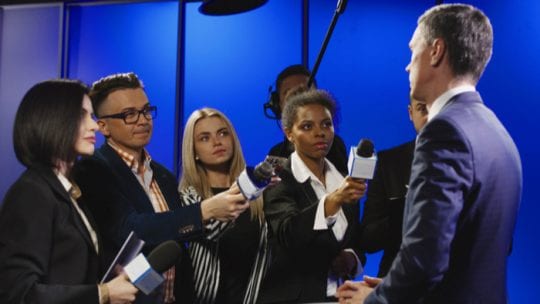 Big group of multiracial journalists with microphones and other technological equipment having interview with middle-aged spokesperson in underlit studio.