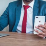 An African-American man dressed in a sharp blue suit with a white shirt and a vibrant red tie, holding an iPhone in his hand. The image focuses on the upper body, showcasing professional attire and the modern device.