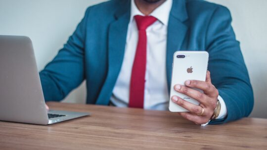 An African-American man dressed in a sharp blue suit with a white shirt and a vibrant red tie, holding an iPhone in his hand. The image focuses on the upper body, showcasing professional attire and the modern device.