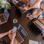 Four coworkers sitting around a table, each using their own laptop while working on a project together. They are focused on their screens, typing and scrolling through documents. The table is cluttered with notebooks, pens, and glasses of water.