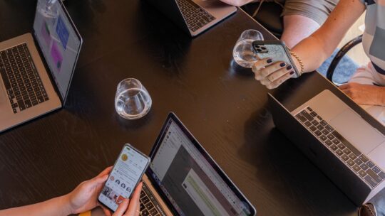 Four coworkers sitting around a table, each using their own laptop while working on a project together. They are focused on their screens, typing and scrolling through documents. The table is cluttered with notebooks, pens, and glasses of water.