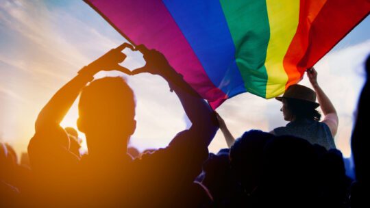 Pride community at a parade with hands raised and the LGBT flag.