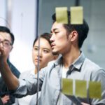 young asian entrepreneur of small company putting a adhesive note on glass in office during team meeting formulating business strategies.