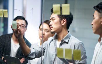 young asian entrepreneur of small company putting a adhesive note on glass in office during team meeting formulating business strategies.