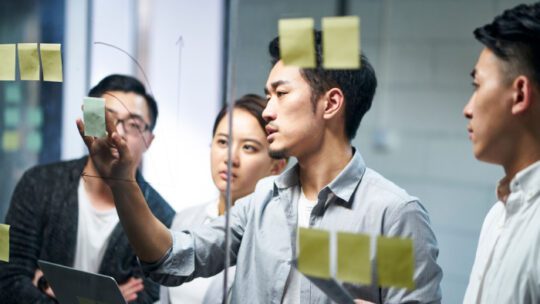 young asian entrepreneur of small company putting a adhesive note on glass in office during team meeting formulating business strategies.