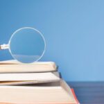 open book with magnifying glass on wooden desk in information library of school or university, concept for education,reading , study, copy space and blue background.