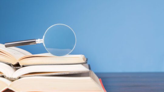 open book with magnifying glass on wooden desk in information library of school or university, concept for education,reading , study, copy space and blue background.