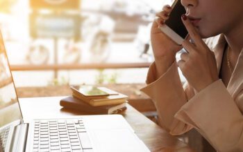 A woman puts her finger up to her lips in a shushing sign, while on the phone, looking at a laptop screen. She is telling the person on the other end that this is important information for an exclusive.