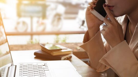 A woman puts her finger up to her lips in a shushing sign, while on the phone, looking at a laptop screen. She is telling the person on the other end that this is important information for an exclusive.