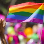 A supporting hand waves in front of a rainbow flag flying on the sidelines of a summer gay pride parade