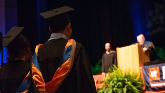 College students dressed in cap and gowns listen to a speech during a graduation ceremony.