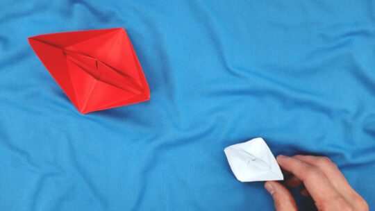 Competition with large corporation and big players concept. Small paper boat facing a large ship.