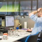 Male journalist working at a computer, putting a hand on his head in frustration, sitting on a chair.