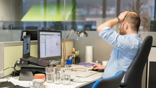 Male journalist working at a computer, putting a hand on his head in frustration, sitting on a chair.