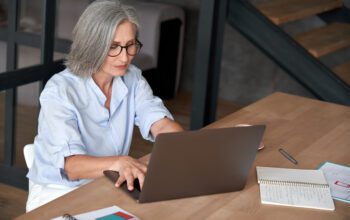 Older mature middle aged business woman employee using laptop typing computer sitting at workplace desk. Senior old lady 60s grey-haired businesswoman entrepreneur or executive working on pc in office