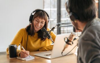 Female podcaster in headphones interviewing man guest using microphone and laptop, writing in notebook, smiling, recording and broadcasting audio podcast in studio. Content creator, blogging