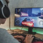 teenager wearing headphones plays online car video game on personal computer. Professional young gamer spends time at home, enjoys gameplay in street racing drift simulator, uses keyboard.