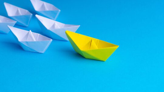 White and yellow paper boat or ship in one direction on blue background, representative of a thought leader engaging the community.