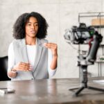 A Black businesswoman engages confidently with a camera, gesturing as if explaining a complex concept during a video blog or webinar session in a modern office environment.