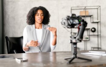 A Black businesswoman engages confidently with a camera, gesturing as if explaining a complex concept during a video blog or webinar session in a modern office environment.