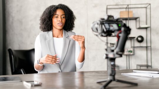 A Black businesswoman engages confidently with a camera, gesturing as if explaining a complex concept during a video blog or webinar session in a modern office environment.