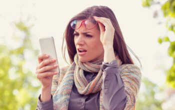 Annoyed upset woman in glasses looking at her smart phone with frustration while walking on a street on an autumn day