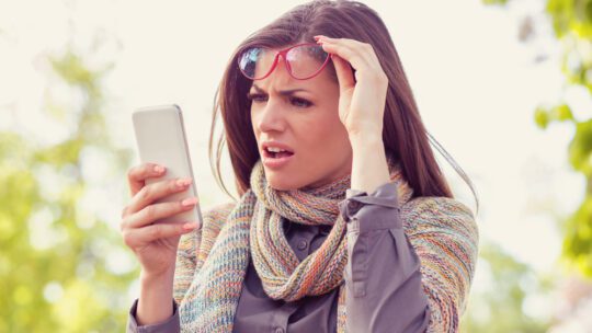 Annoyed upset woman in glasses looking at her smart phone with frustration while walking on a street on an autumn day