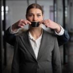 Woman in a suit stands in an office with a serious expression, as a man behind her silences her with black tape over her mouth. corporate silence