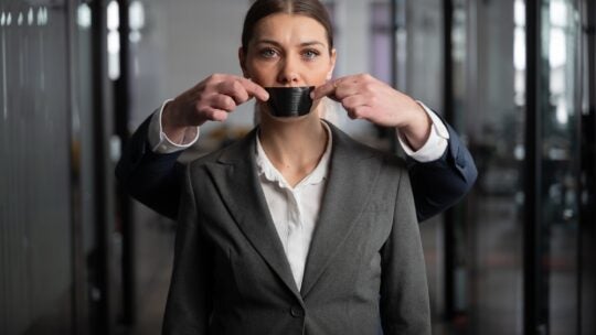Woman in a suit stands in an office with a serious expression, as a man behind her silences her with black tape over her mouth. corporate silence