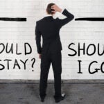 Rear view of confused businessman looking at should I stay or go text and arrows going 2 ways to represent staying or leaving a job