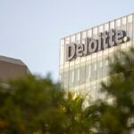 This image features the top of the Deloitte building, with the company's name prominently displayed in large letters on the glass facade.