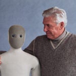 elderly man squeezing the Neo robot around his shoulders like a hug. this relates to the product mentioned in this bad coverage article.