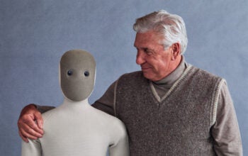 elderly man squeezing the Neo robot around his shoulders like a hug. this relates to the product mentioned in this bad coverage article.