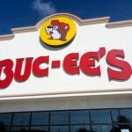A Buc‑ee’s roadside convenience superstore displays its signature beaver‑mascot logo and bold red lettering on a light‑colored façade along a Texas travel corridor, reflecting the chain’s oversized retail footprint and its role in regional highway culture. Houston, TX