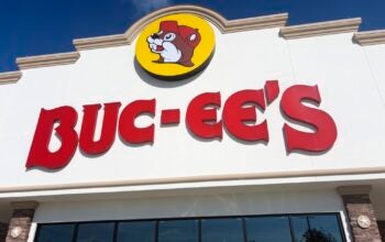A Buc‑ee’s roadside convenience superstore displays its signature beaver‑mascot logo and bold red lettering on a light‑colored façade along a Texas travel corridor, reflecting the chain’s oversized retail footprint and its role in regional highway culture. Houston, TX