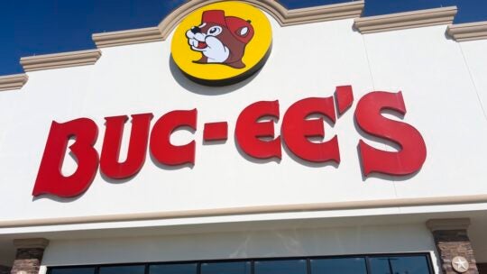 A Buc‑ee’s roadside convenience superstore displays its signature beaver‑mascot logo and bold red lettering on a light‑colored façade along a Texas travel corridor, reflecting the chain’s oversized retail footprint and its role in regional highway culture. Houston, TX