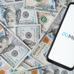 Meta new facebook company logo on smartphone screen, social network mobile application. Top view gadget lying on money background of one hundred dollar bills.